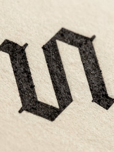Macro shot of deep black ink pressed onto textured warm cream paper, showing the physical texture of typographic ligatures under soft natural daylight.