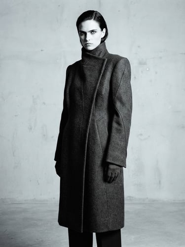 High-contrast minimalist fashion photography, model in structural charcoal wool coat, sharp directional studio lighting, desaturated cool tones, negative space