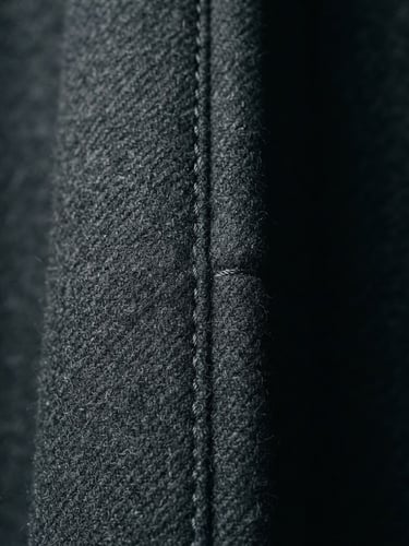 A close-up of a structural garment seam on a minimalist charcoal coat, sharp directional studio lighting, high contrast, desaturated cool tones