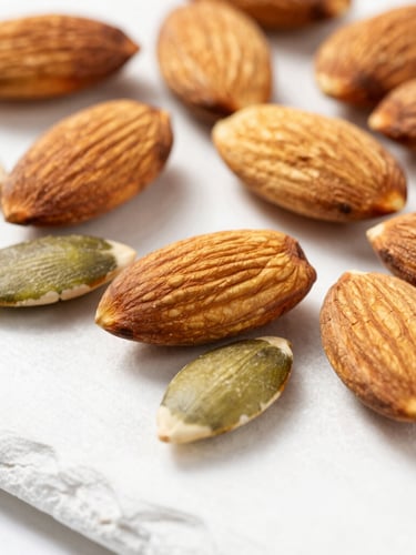 Macro close-up of premium raw almonds and clean pumpkin seeds scattered on a white slate surface, bright natural light, sharp focus, modern athletic aesthetic