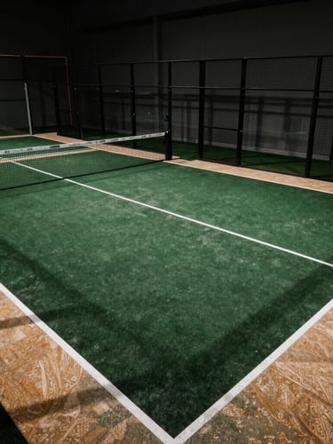 Dramatic wide-angle architectural photography of the indoor padel courts showing their structural scale, deep forest green courts with warm travertine borders, high-contrast shadows.
