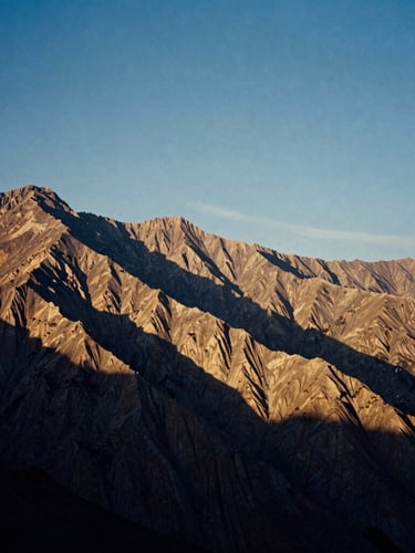 Documentary 35mm film photo of vast, wind-swept Himalayan cliffs at golden hour, high contrast, deep indigo shadows and warm parchment light, expansive sky.