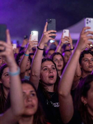 Close-up portrait frame of a crowd mid-cheer at a festival, faces lit simultaneously by stage wash in electric violet and the glow of phone screens held aloft, physical and digital worlds visible in the same moment, high-contrast lighting, tight emotional framing