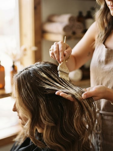 Warm editorial close-up of a stylist gently applying a creamy botanical hair treatment mask, soft natural window light, rich cream and coffee tones, shallow depth of field, 35mm.