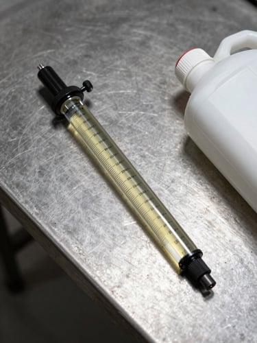 Overhead studio shot of a refractometer resting on a steel maintenance bench beside an open lubricant container, diffused strobe lighting showing true fluid color and instrument scale detail, concrete surface beneath, right-edge composition