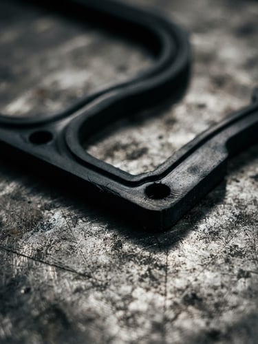 Extreme close-up macro photography of a finished black elastomer gasket showing clean die-cut edges on a steel workbench, dramatic industrial workshop lighting, sharp focus