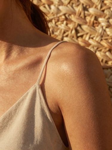 Close-up of sun-kissed skin and raw linen fabric, golden hour light filtering through woven straw, warm organic textures.