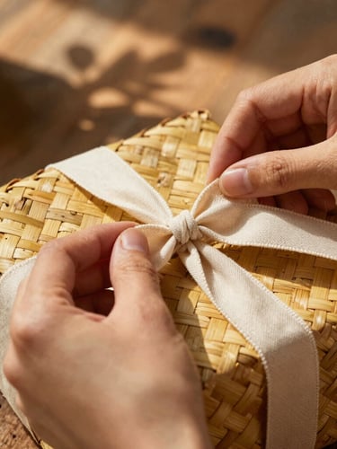 Macro close-up of hands tying a soft linen ribbon around a woven straw gift basket, warm golden hour sunlight filtering through leaves, dapple shadows on a rustic wooden table.