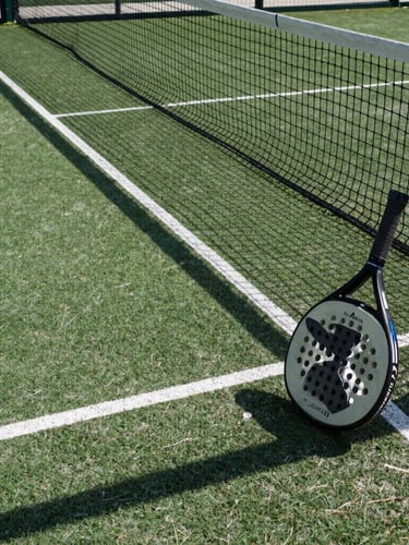 Dynamic low-angle shot of a modern outdoor padel court, sharp morning shadows on the deep green surface, crisp white lines, racket resting against the net.