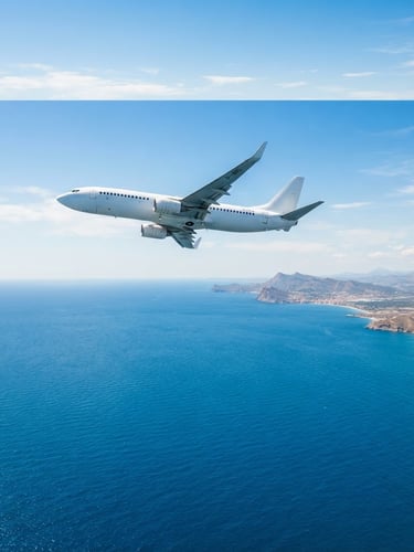 Bright editorial photo of a commercial airplane flying high above the bright blue Mediterranean Sea towards the coast of Europe, sunny daylight, 35mm.