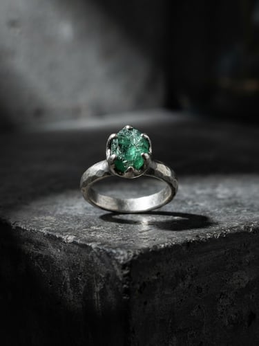Fine art jewelry photography, a raw silver ring with an uncut emerald on a dark concrete pedestal, dramatic gallery spotlight, deep architectural shadows, Hasselblad 80mm