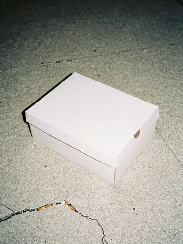 Harsh direct flash photography of a sneaker box resting on cracked raw concrete, gloomy daylight, high contrast desaturated tones