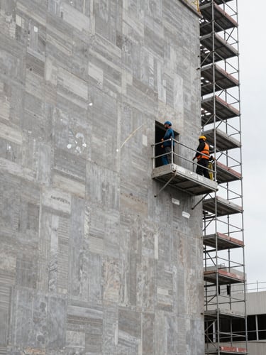 Wide environmental shot of a building facade under active renovation, scaffolding on the right third, workers on platform mid-task, morning overcast light, cool neutral tones, no posed figures