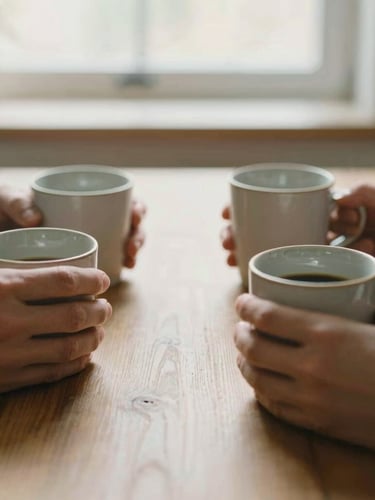 Warm candid close-up of hands holding ceramic coffee mugs around a wooden table, soft morning daylight streaming through a window, shallow depth of field
