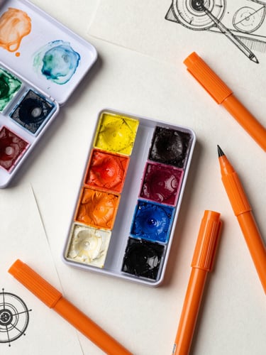 Overhead flat-lay of a graphic designer's desk with watercolor paint pans, vibrant orange alcohol markers, and textured paper sketches under soft natural daylight