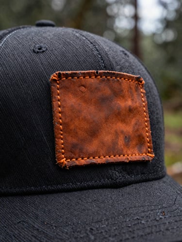 Macro shot of a rugged, weathered leather patch stitched onto a dark trucker hat, overcast outdoor forest background, shallow depth of field