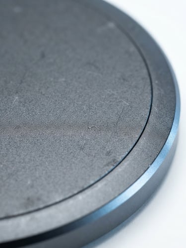 Macro shot of an anodized aluminum wireless charger casing, bright high-key studio lighting with soft cool-blue ambient bounce and subtle rim highlights, extreme close-up focusing on material textures, clean white backdrop