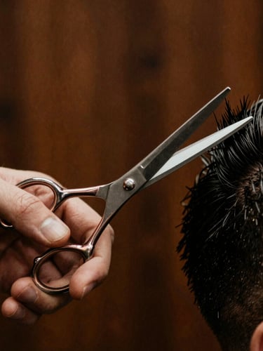 Close-up of a barber's hands using vintage steel scissors to style hair, warm low-key directional lighting, shallow depth of field, dark wood background.