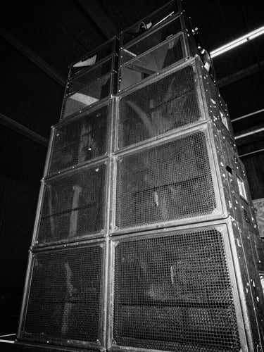 Low-angle wide shot of a towering, gritty warehouse speaker stack, high-contrast black and white, heavy film grain, strobe-lit shadows.