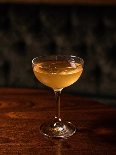 Close-up of a perfectly crafted cocktail in a premium glass, resting on a dark wood table with a blurred velvet texture in the background, dramatic chiaroscuro lighting, warm amber tones, shallow depth of field, 35mm film.