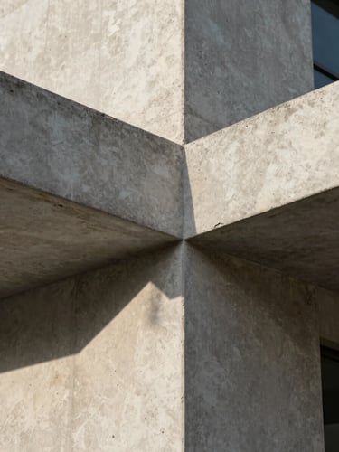 A close-up architectural detail of raw board-formed concrete intersecting with honed travertine under low-angle morning sunlight, showing texture and shadow.