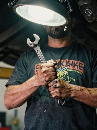 Close-up of a heavy-weight black graphic t-shirt worn by a mechanic under a car lift, hands stained with grease holding a wrench, harsh overhead shop light, gritty 35mm film texture