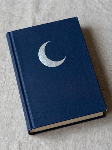 Close portrait of a hardbound book lying on aged linen fabric, cover facing up — deep indigo cloth binding with a pale silver crescent moon embossed at center, soft candlelight raking across the texture of the cover from lower left, cool shadow on the right third, no text visible, intimate cabinet-of-curiosities framing