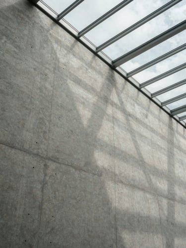 Wide-angle architectural crop of a concrete wall meeting a glass ceiling, dramatic light-and-shadow play, overcast daylight.