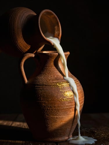 Dramatic side light shining on an ancient textured clay jug pouring milk, deep chiaroscuro shadows, gold glints.