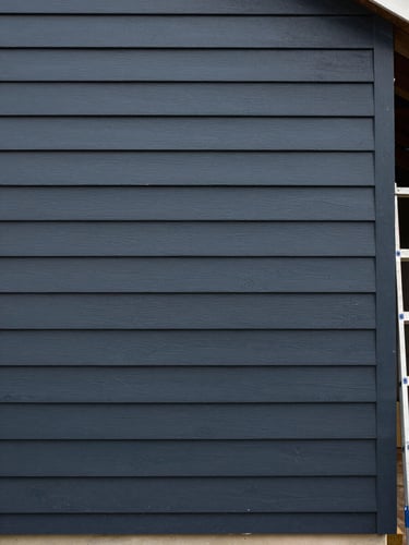 Straight-on documentary shot of a freshly painted exterior clapboard siding wall, deep charcoal paint showing even sheen, overcast daylight revealing true color without glare, ladder edge visible at one side indicating active job site