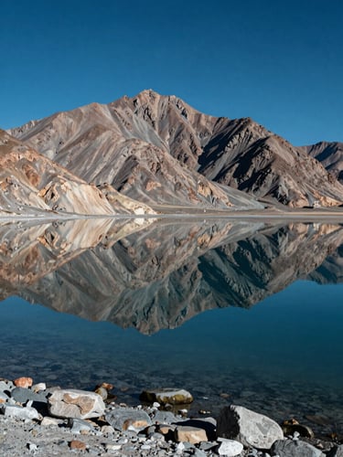 Deep blue high-altitude salt lake Pangong Tso reflecting sharp barren mountains under a clear sky, stone shoreline in foreground