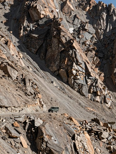 A high-altitude mountain pass in Ladakh, a single rugged 4x4 vehicle dwarfed by massive slate-colored cliffs under harsh sunlight