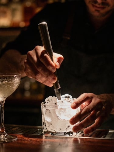 Intimate portrait crop of a bartender carefully carving clear ice under a warm, low-key spotlight, condensation-beaded cocktail glasses nearby, moody atmosphere.