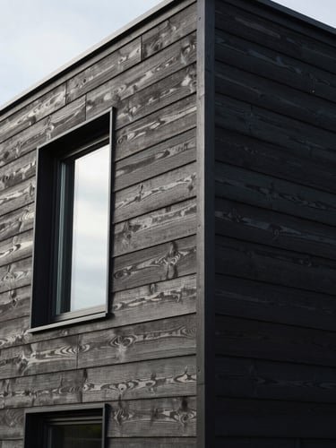 Close-up architectural detail of a contemporary UK house extension facade, board-on-board dark timber cladding with slim aluminium window frame catching diffuse overcast daylight, precise shadow lines at panel joints, no people, wide environmental framing showing the full wall elevation