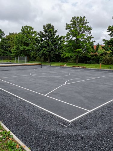Architectural outdoor photography of a large sports court under construction, showing the technical base preparation and gravel compaction, soft overcast daylight, deep grays, wide-angle lens, sharp details.