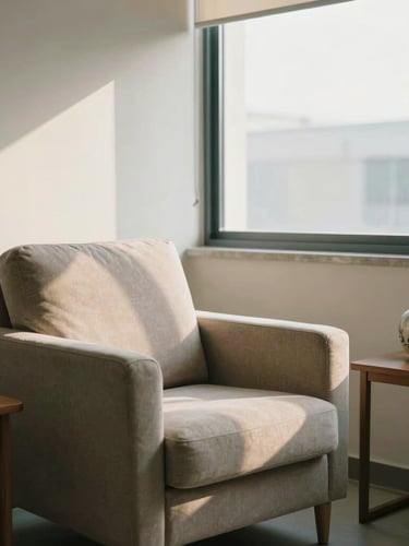 A quiet, sunlit clinical consultation room with a soft fabric armchair, a small wooden side table with a ceramic vase, warm morning light streaming through a large window, peaceful and dignified atmosphere.