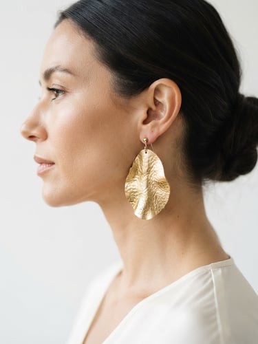 Close-up profile of an elegant woman wearing a large gold-textured leather earring, soft natural daylight, minimalist white background, editorial crop
