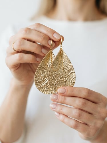Close-up of elegant hands holding a pair of large, gold-textured leather earrings, bright natural daylight, minimalist white background