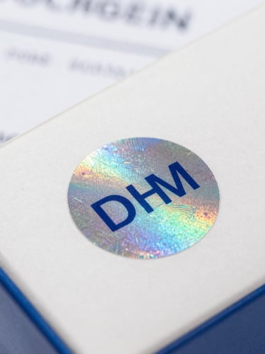 Close-up macro shot of a genuine OEM holographic seal on a product box, with official looking paperwork partially visible in the background. Bright, neutral commercial lighting.