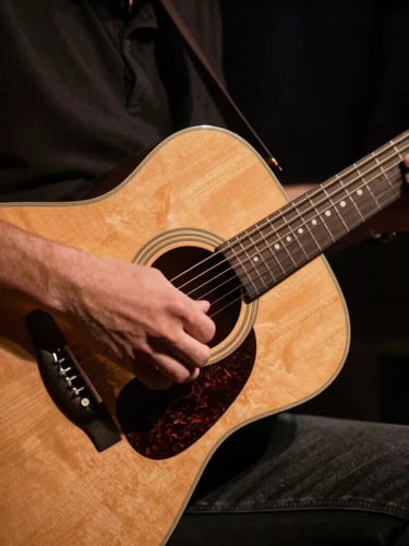 Tight close-up of hands playing an acoustic guitar on a dark stage, warm amber spotlight catching the wood grain and strings, moody atmosphere.