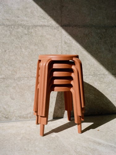 Low-angle architectural photograph of terracotta-toned stackable resin stools against a raw board-formed concrete wall, sharp geometric midday shadows, 35mm
