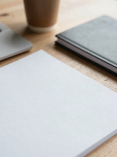 Close-up of a wooden desk with research papers, a modern notebook, and a coffee cup in soft morning light, minimalist academic aesthetic