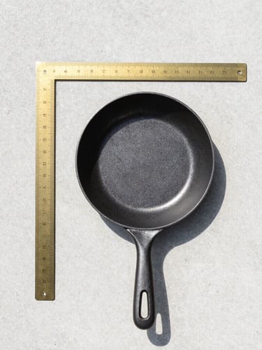 Orthographic top-down view of a single heavy brass ruler and a matte black cast-iron pan arranged geometrically on a flat concrete surface, bright high-key daylight, crisp short shadows