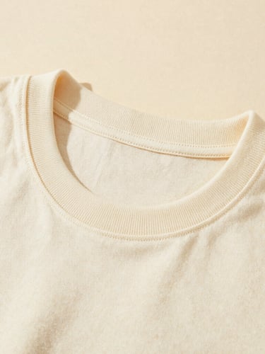 Close-up of a heavyweight cream cotton T-shirt collar, showing clean single-needle stitching, warm directional window light, rich tactile texture