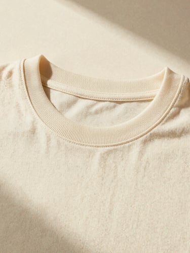 Close-up of a heavyweight cream cotton T-shirt laid flat, showing the thick single-needle stitched collar and clean shoulder seams. Warm directional studio light, soft shadows, neutral cream background.