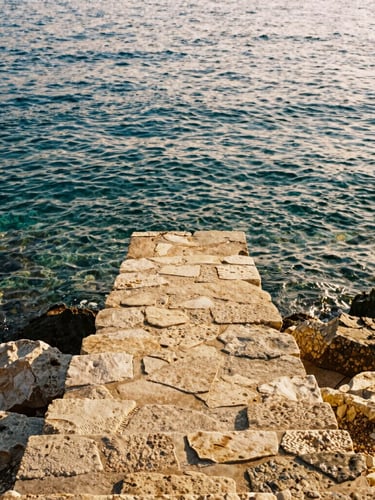 A sun-drenched stone path leading to the Adriatic sea in Dubrovnik, warm golden-hour light reflecting on clear turquoise water, 35mm film aesthetic.