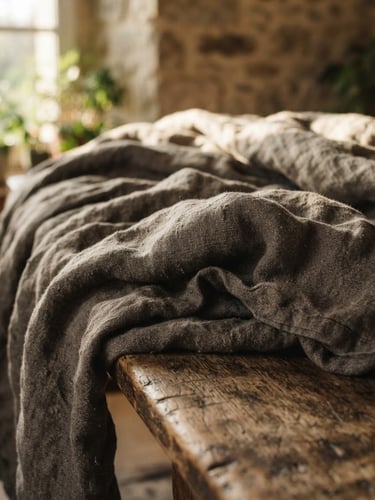 A close-up of a rumpled heavy-weight linen quilt draped over a worn wooden bench, soft morning light casting deep shadows on the textured weave.