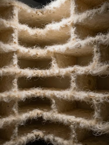 Macro photography of dense nonwoven fiber loft, dramatic side-lighting emphasizing the layered structural density and acoustic chambers, architectural framing.