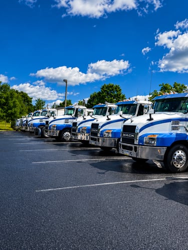 Fleet of blue and white Mack commercial trucks parked in a row under a bright blue sky.