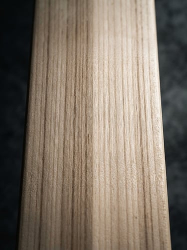 Close-up of a premium cricket bat face showing tight, straight grain lines on a dark concrete studio background with dramatic side lighting.